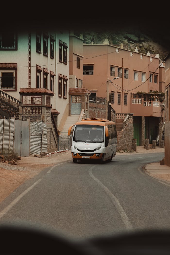 A bus travels down a narrow, winding road in a rustic village with earthy-toned buildings.