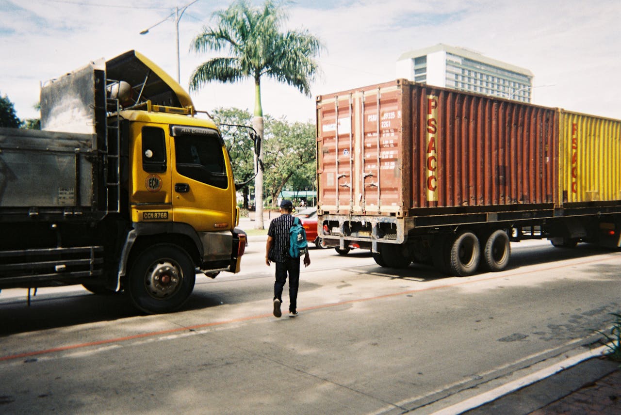 A man walks across a bustling street between two large trucks, under a sunny sky.